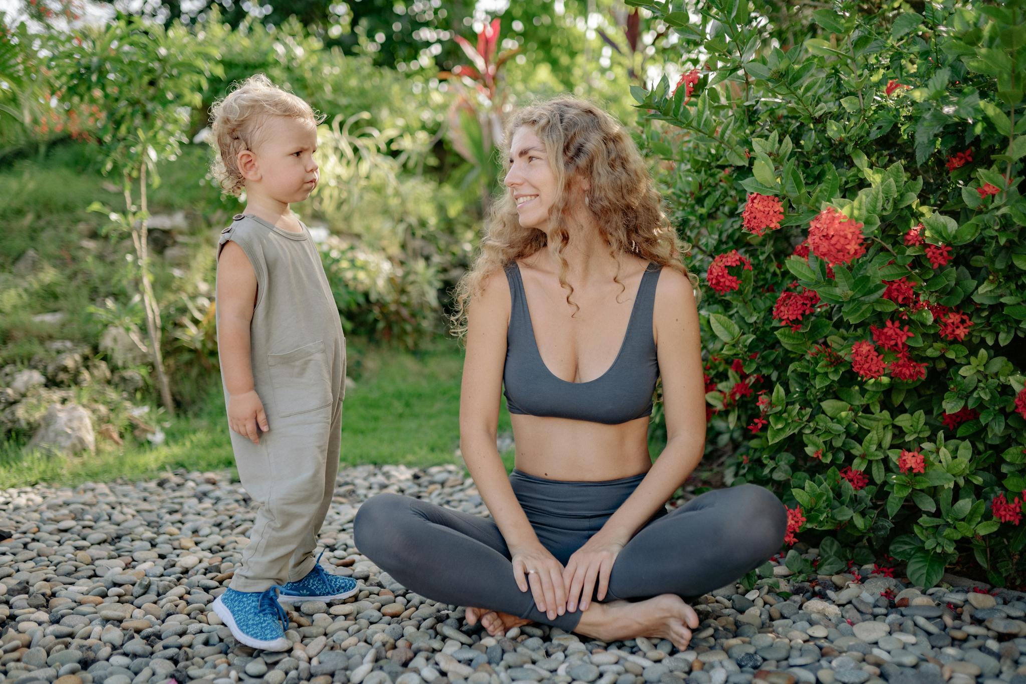 Positive barefoot woman in sports clothes sitting with crossed legs while looking at adorable kid standing on stones in colorful park in summer