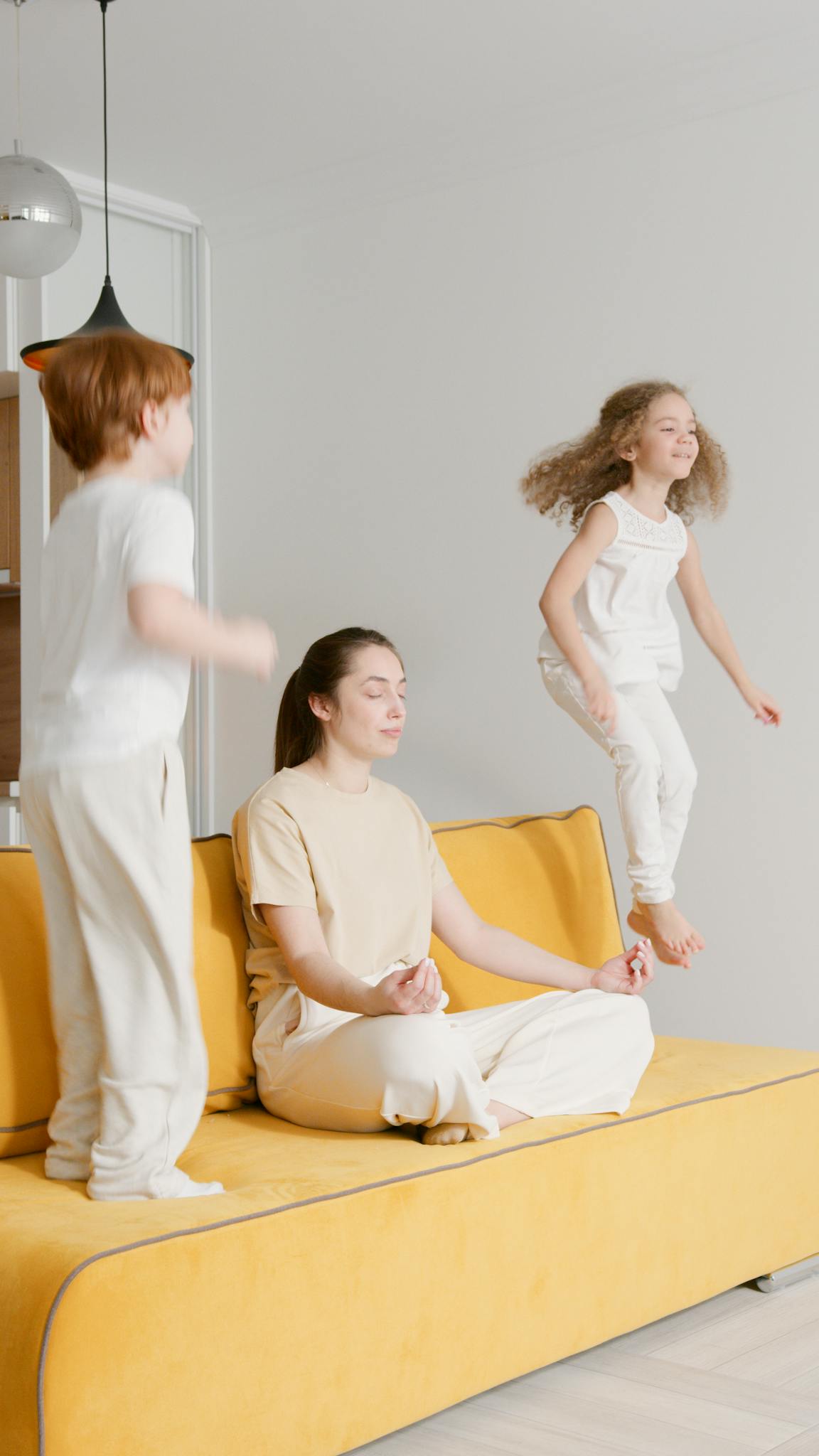 A serene woman meditates on a sofa while two children energetically jump beside her.
